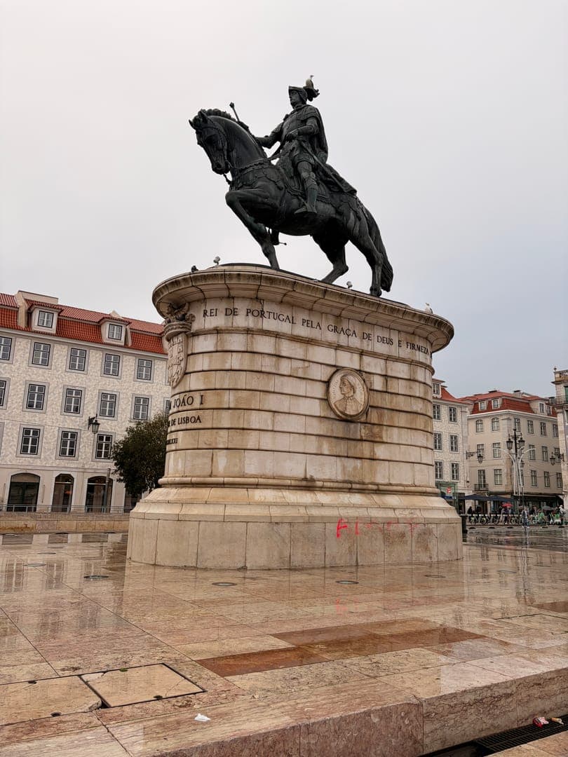 This striking image captures the equestrian statue of King João I in Lisbon, Portugal, under a cloudy sky. The statue, prominently featuring the inscription 'REI DE PORTUGAL PELA GRAÇA DE DEUS E FIRMEZA,' stands on a grand stone pedestal, with historic buildings visible in the background. The wet, reflective ground adds a dramatic touch, enhancing the monument's imposing presence.