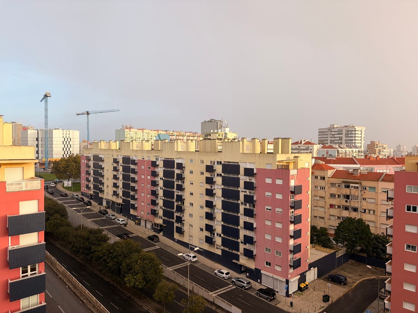 A vibrant cityscape unfolds under a soft, overcast sky, showcasing a mix of residential buildings with striking color palettes. The foreground features a red and black building, while a prominent cream and pink building with dark blue balconies dominates the midground, flanked by a busy street with parked cars and lush trees. In the distance, construction cranes rise above the urban sprawl, hinting at ongoing development and a dynamic city life.