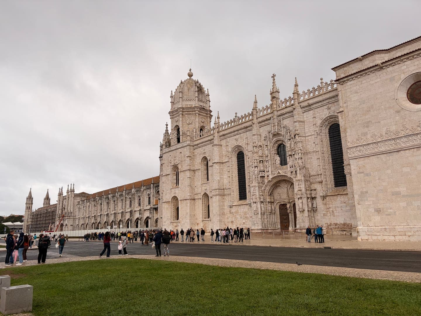 The Jerónimos Monastery in Lisbon, Portugal, stands majestically under a cloudy sky, showcasing its intricate Manueline architecture. A crowd of people gathers outside the grand entrance, admiring the detailed carvings and towering spires of this historic landmark. The lush green grass in the foreground adds a touch of natural beauty to the scene, highlighting the monastery's impressive scale and timeless elegance.
