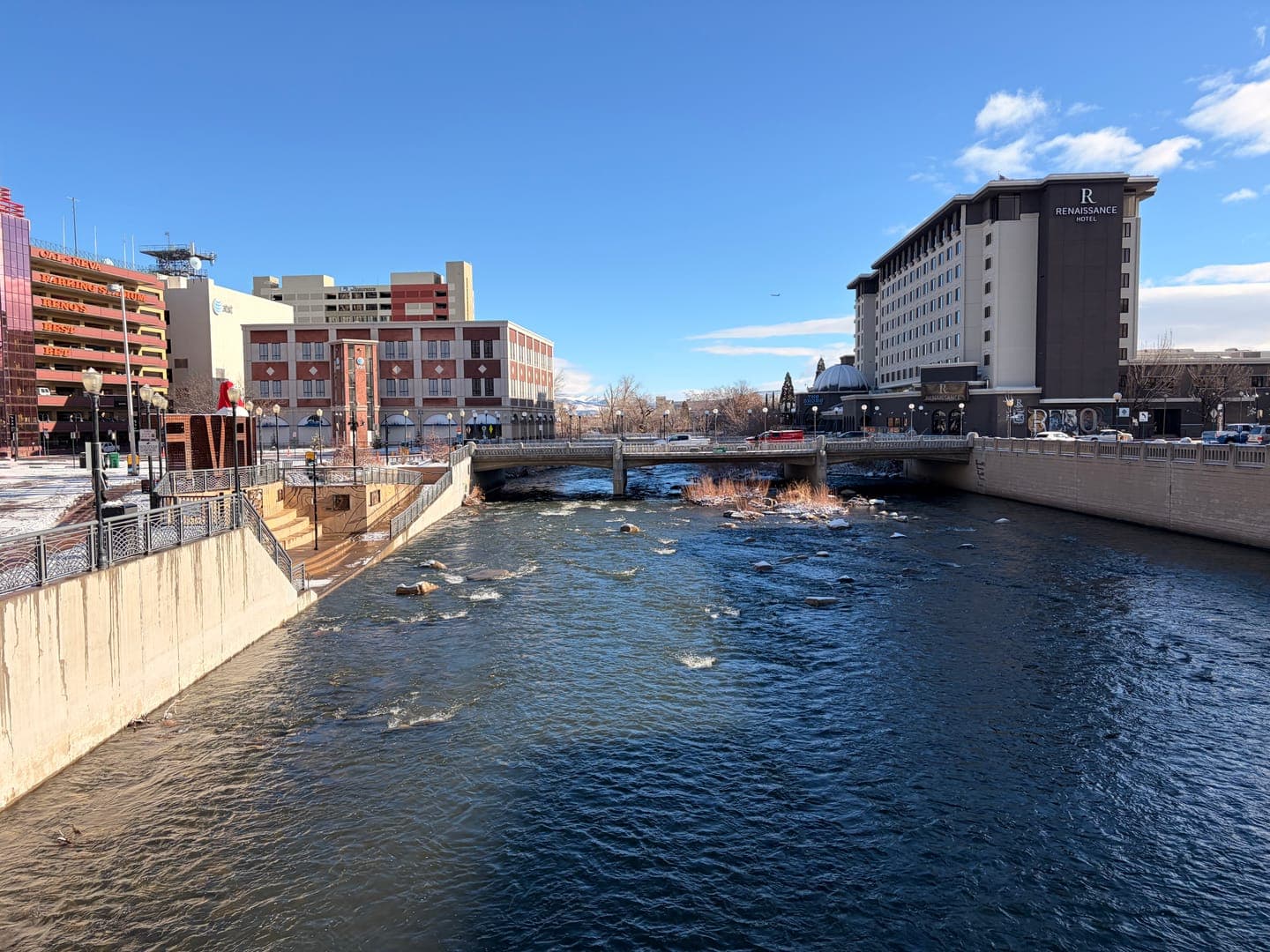 A vibrant cityscape unfolds along the Truckee River in Reno, Nevada, under a clear blue sky. The Renaissance Hotel stands prominently on the right bank, its name clearly visible, while other buildings and bridges line the river's edge. The flowing water, with hints of snow and ice on the banks, adds a dynamic element to this urban winter scene.