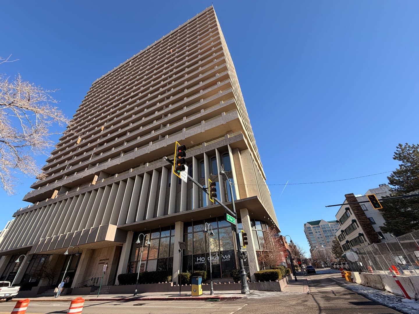 This low-angle shot captures a towering, modern high-rise building with numerous balconies under a clear blue sky. The building's base features large windows, revealing a business named 'Ho kava' and 'Cigar Lounge' inside, while traffic lights and street signs are visible in the foreground. The bright sunlight and crisp blue sky create a vibrant, urban atmosphere, with a hint of winter from the bare tree branches and snow on the ground.