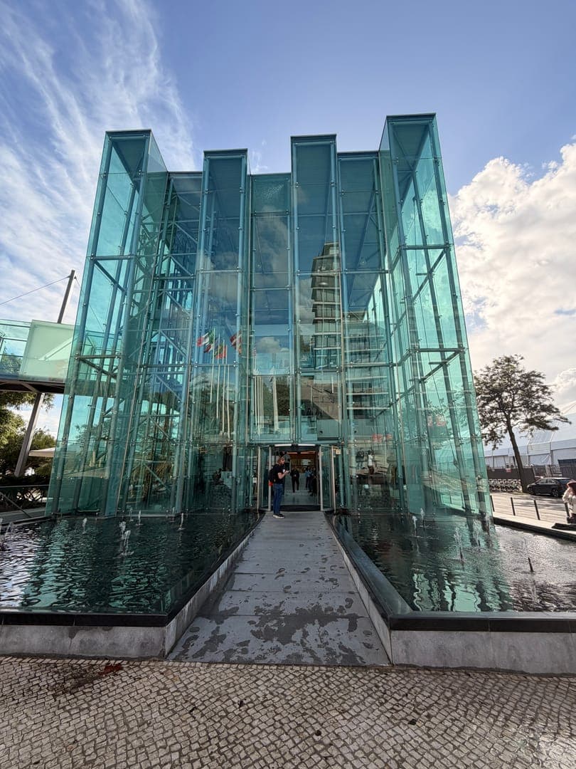 This striking image captures a modern glass building, its transparent walls reflecting the vibrant blue sky and fluffy white clouds above. A serene water feature with gentle fountains surrounds the entrance, creating a tranquil atmosphere. The architectural design, with its clean lines and reflective surfaces, offers a captivating blend of contemporary style and natural beauty.
