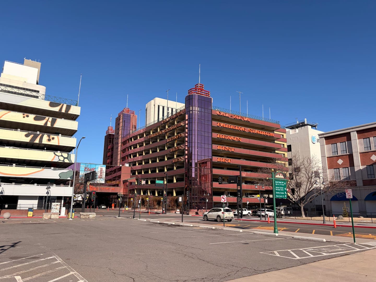 This vibrant street scene captures the unique architecture of downtown Reno, Nevada, under a clear blue sky. The prominent red-orange parking garage, emblazoned with 'CAL-NEVA,' 'RENO'S,' 'BIGGEST,' and 'BEST,' stands out against the lighter-colored buildings, including one with an AT&T logo. The image conveys a bright, urban atmosphere with a mix of modern and older structures, hinting at the city's lively character.