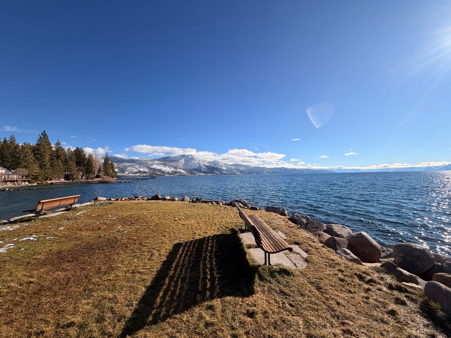 Two wooden benches sit on a grassy bank overlooking a vast, sparkling lake under a clear blue sky. In the distance, snow-capped mountains rise majestically, framed by a line of evergreen trees and a few scattered clouds. The bright sun casts long shadows from the benches, creating a serene and inviting scene perfect for quiet contemplation.