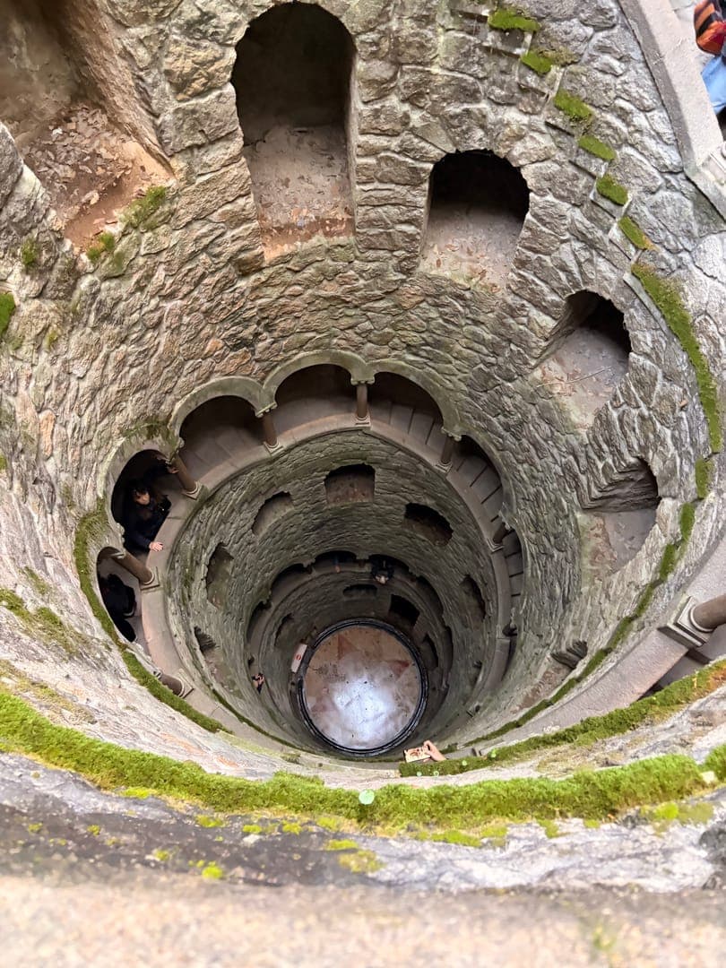 Step into the mysterious depths of the Initiation Well at Quinta da Regaleira, Portugal. This captivating shot looks down into the spiral staircase, revealing its intricate stone architecture, moss-covered edges, and the intriguing circular pattern at its base. The well's unique design and the play of light and shadow create an atmosphere of ancient wonder and hidden secrets.
