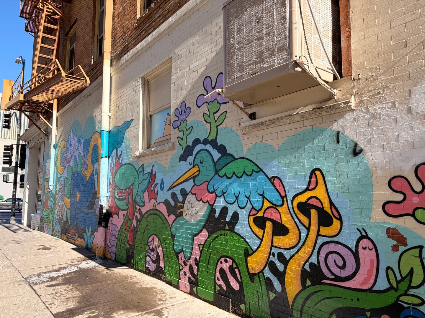 This vibrant street art transforms a plain brick wall into a whimsical wonderland. Featuring colorful cartoon-like creatures, including a blue bird, snails, and mushrooms, the mural brings a playful and imaginative mood to the urban landscape. The detailed artwork, set against a light blue and white brick background, creates a striking contrast with the industrial fire escape and air conditioning unit above.