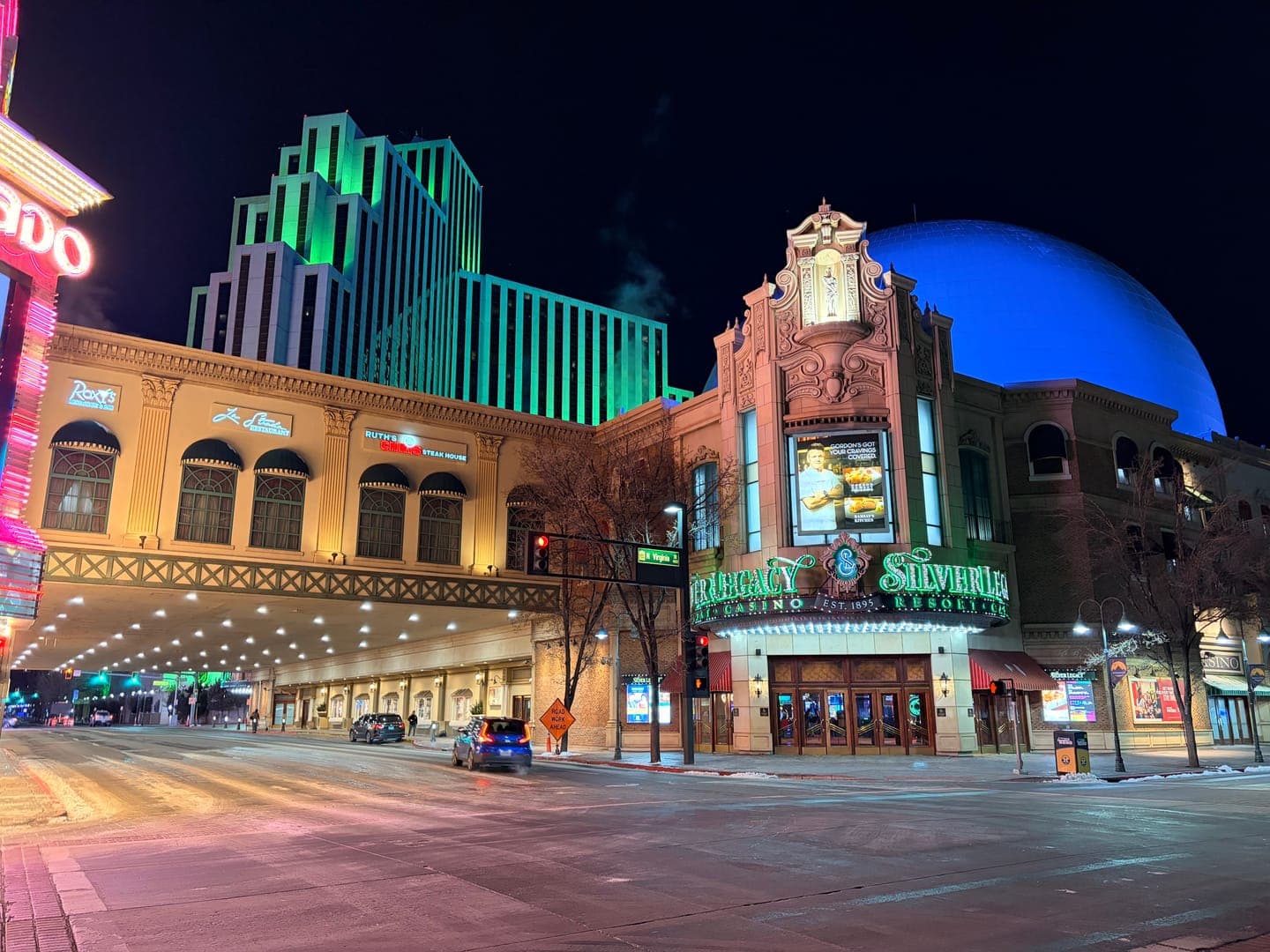 The Silver Legacy Resort Casino in Reno, Nevada, glows vibrantly at night, with its ornate facade and a prominent sign for 'Silver Legacy Resort Casino' illuminated in green. Behind it, a modern skyscraper also shines with green lights, while a large blue dome adds a striking contrast to the dark sky. The street below is quiet with a few cars, reflecting the colorful lights of the buildings and creating a captivating urban night scene.