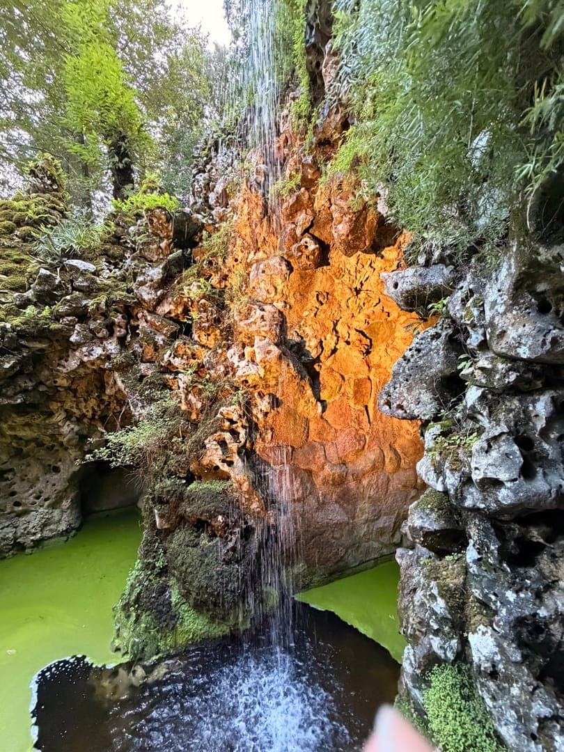 A vibrant waterfall cascades down a moss-covered, orange-hued rock face into a pool of bright green water. Lush greenery and trees frame the top of the scene, adding to the natural, enchanting atmosphere. The striking contrast between the warm rock, cool water, and verdant foliage creates a captivating and serene natural landscape.