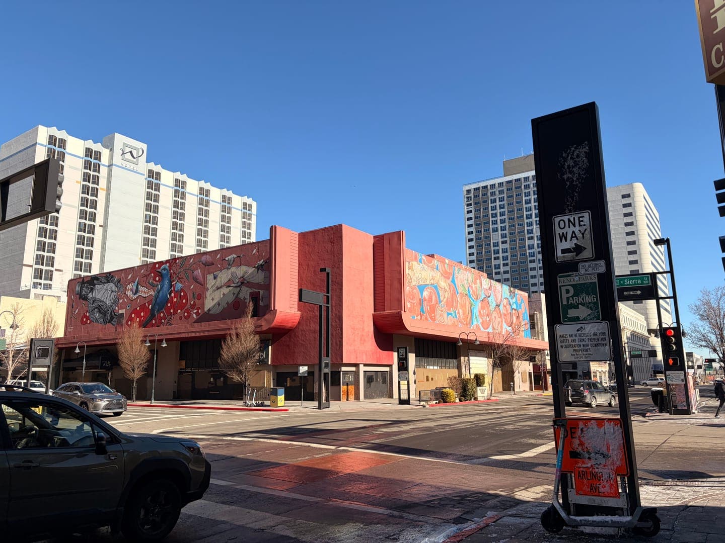 This vibrant street scene captures a colorful mural adorning a red building, with tall white high-rises towering in the background under a clear blue sky. A 'ONE WAY' sign with an arrow pointing right and a 'P PARKING' sign are prominently displayed on a black pole in the foreground, adding to the urban feel. The bright sunlight casts sharp shadows, highlighting the architectural details and the lively street art.