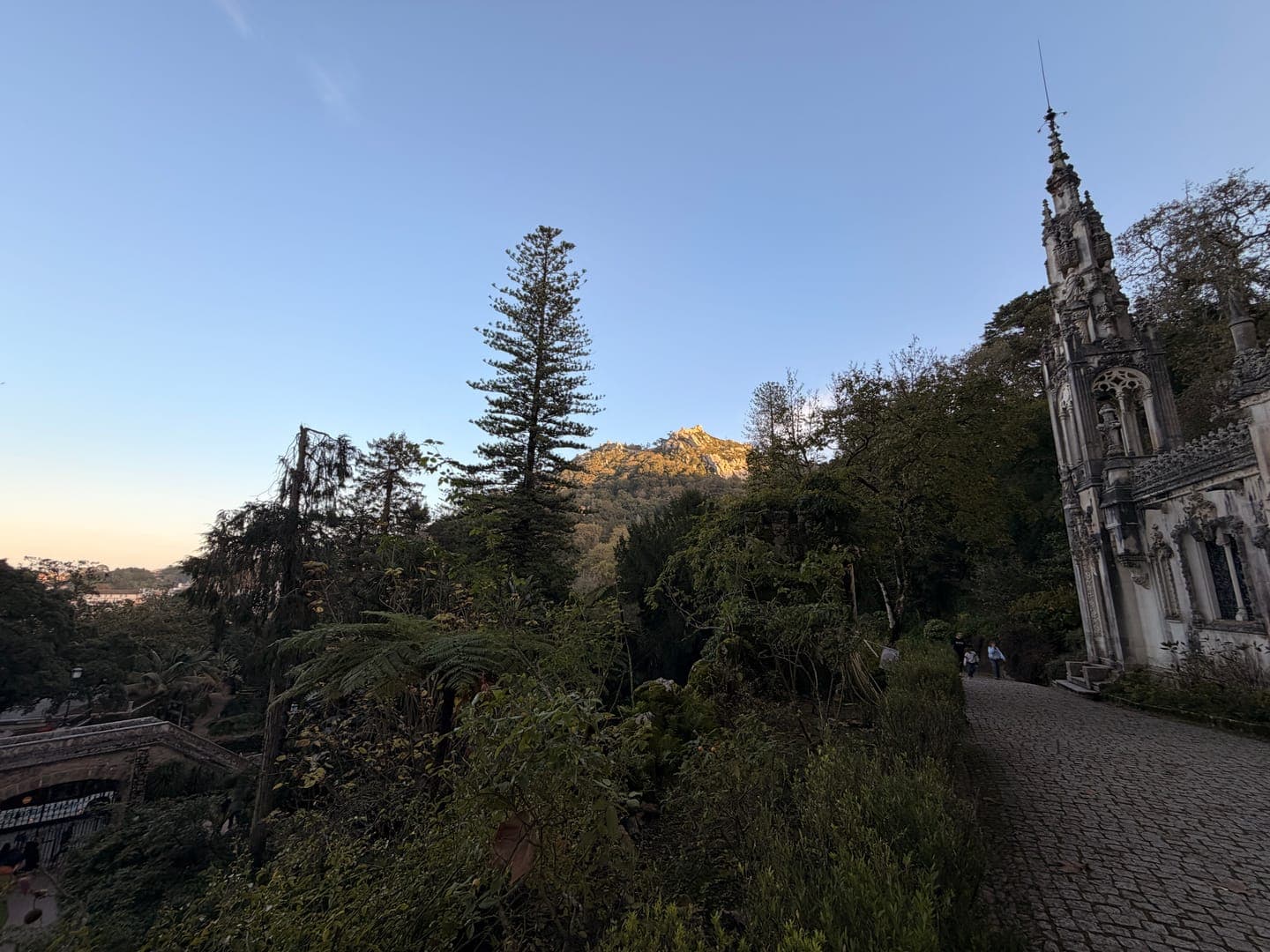A majestic Gothic-style tower rises on the right, its intricate details hinting at a rich history, while a winding cobblestone path leads towards it. Lush green foliage and towering trees frame the scene, with a glimpse of distant mountains under a clear blue sky, creating a serene and enchanting atmosphere. The soft light of what appears to be late afternoon or early morning bathes the landscape, adding to the tranquil beauty of this architectural and natural wonder.